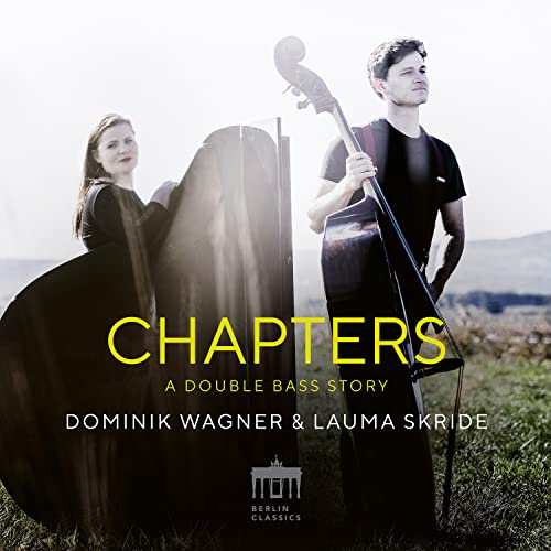 Two musicians with double basses in a sunlit, grassy landscape.