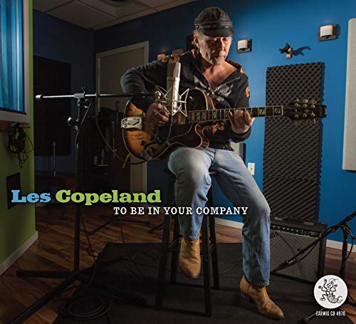 "Man in recording studio playing guitar, seated on stool, with text overlay."