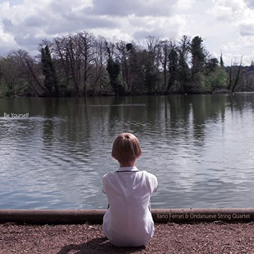 Child sits by a lake, facing trees under a cloudy sky.