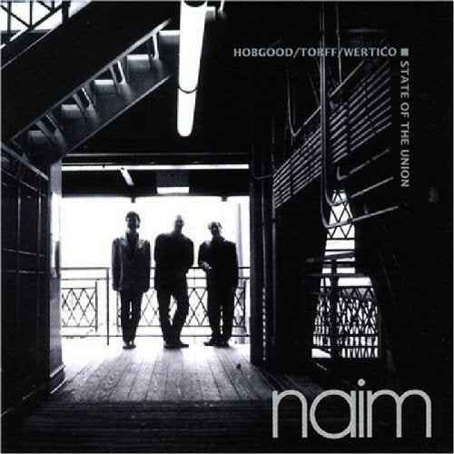 Black and white image of three men standing on a wooden balcony, with "naim" at the bottom.