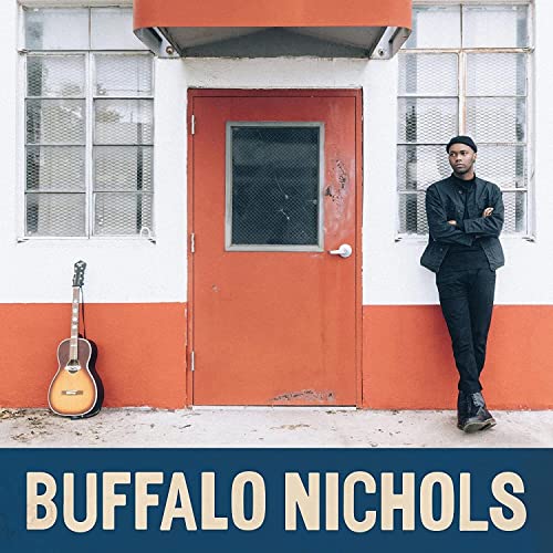 Man with guitar stands before a red door and windowed wall.