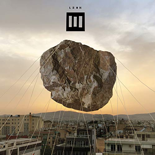 A large rock suspended by cables over a cityscape at sunset.