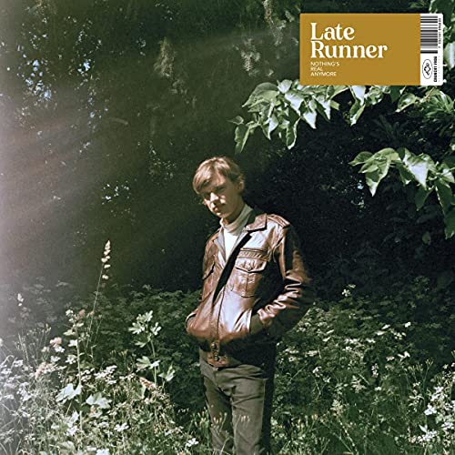 Man in a brown jacket stands in a field of greenery.