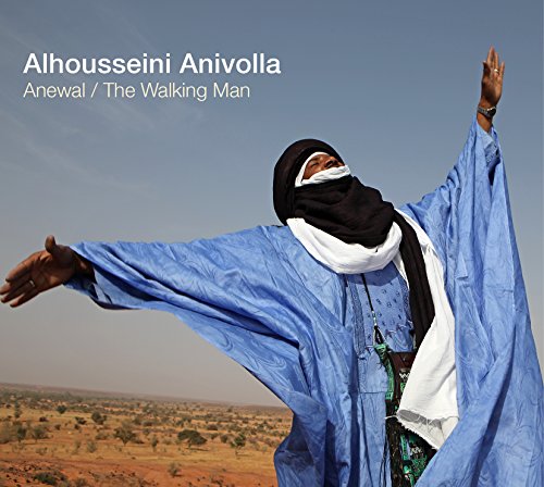 Man in blue robe and black headscarf stands with arms raised against a desert landscape.