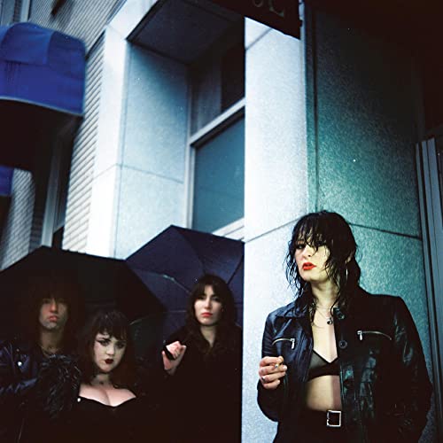 Four musicians with dark hair pose outdoors in front of a building.