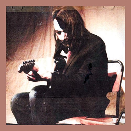 A long-haired man in a dark suit playing an electric guitar while sitting down.