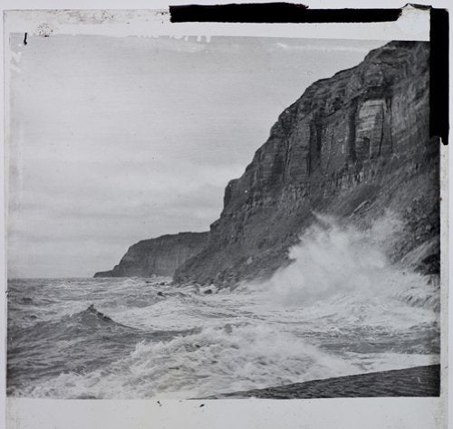 "Black and white photograph of a rugged cliffside with crashing waves."
