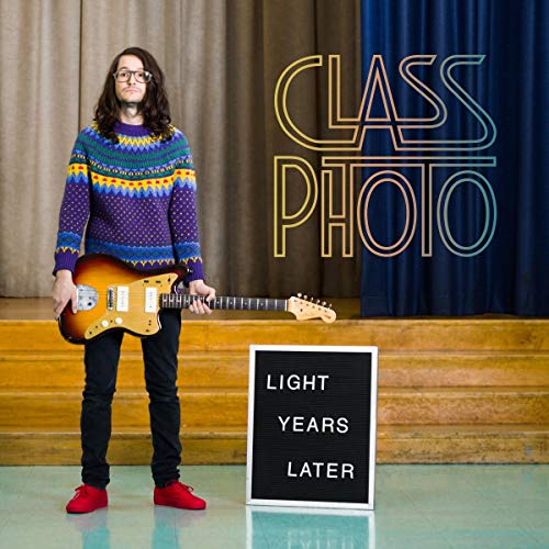 "Man holding guitar stands beside a sign reading 'Light Years Later' on a stage."