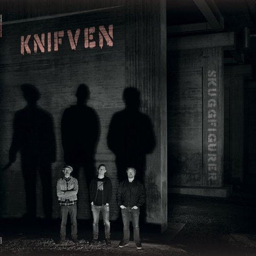 "Three men stand before a wall with their shadows looming large behind them."