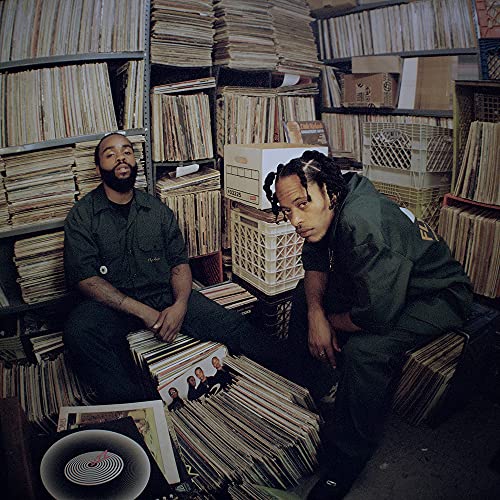 Two musicians sit amidst stacks of vinyl records in a store.