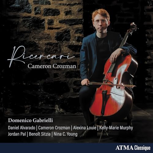 Cellist Cameron Crozman poses with his instrument against a stone wall.