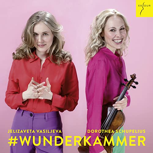 Two women, one holding a violin, posing against a pink background.