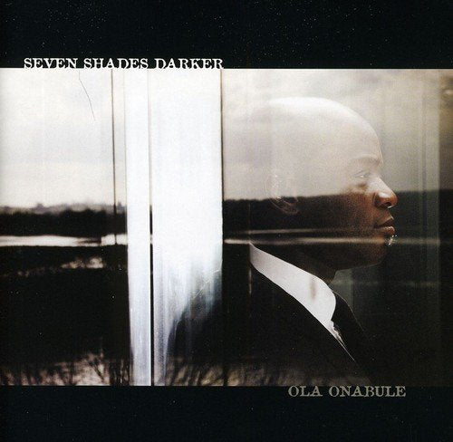 A man in a suit is reflected in a window with the title "Seven Shades Darker".
