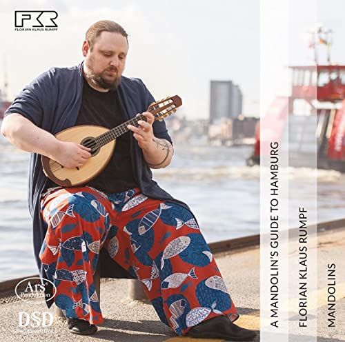 A man playing a mandolin, wearing patterned fish trousers, sitting by a harbor.
