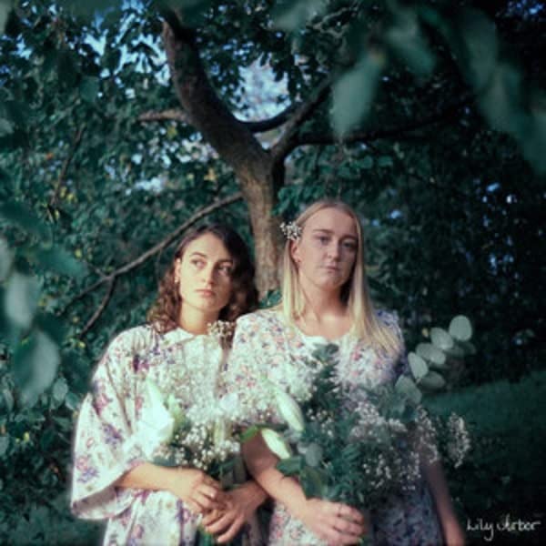 Two women in floral dresses holding bouquets of white flowers under a leafy tree.