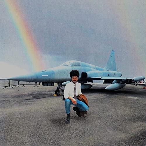 Man sitting near a fighter jet under a rainbow-filled sky.