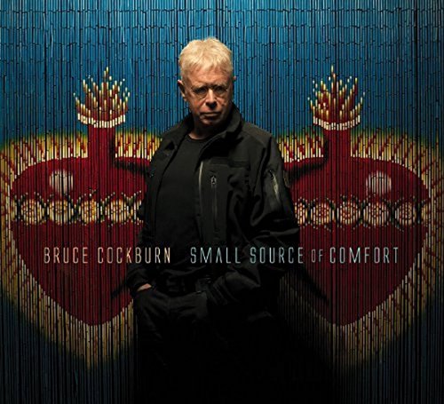 Bruce Cockburn stands before a red heart-shaped tapestry with gold accents.