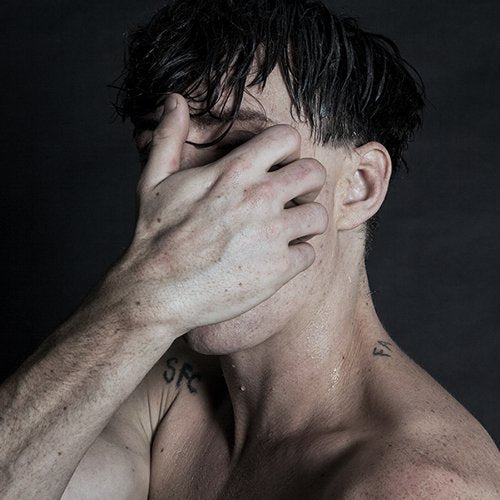 Man with dark hair covers his face with his hand, showing tattoos on his arm and shoulder.