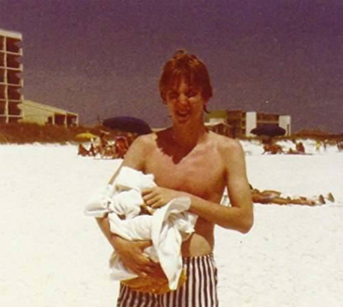 "Man holding baby on a beach, wearing striped shorts, with buildings in the background."