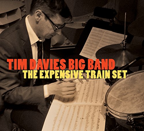"Album cover with man in suit writing on sheet music beside a drum set."