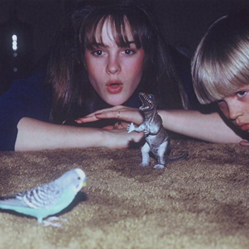 "Two people and a parakeet interacting with a toy dinosaur on a carpet."