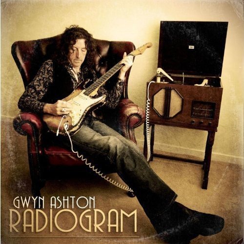 Man sits in chair, playing electric guitar beside an old-fashioned radio.