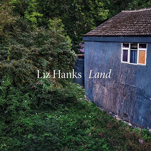 Blue shed with a window, overgrown greenery, album cover for Liz Hanks’ “Land”.