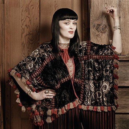 Woman in ornate shawl and striped dress stands against a wooden wall, hand on door frame.