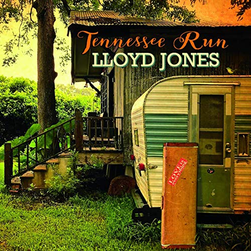 A vintage camper sits next to a wooden building in a grassy, tree-filled area.