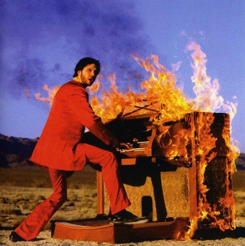 Man in red suit plays a burning piano outdoors, surrounded by flames and smoke.