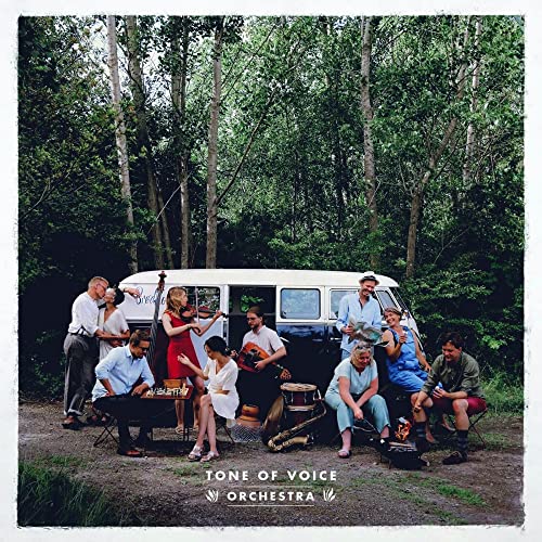 Orchestra group portrait outdoors with instruments, van, and forest backdrop.