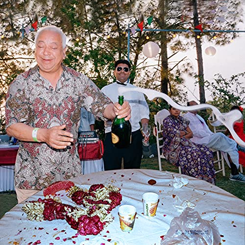 An older man in a patterned shirt pops a champagne bottle at an outdoor celebration.
