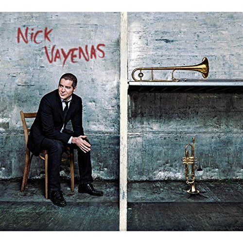 Man in black suit sits on wooden chair next to two brass trumpets on a shelf.