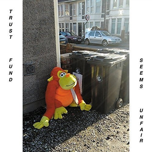 A stuffed monkey in orange and green sits beside a trash bin outdoors.