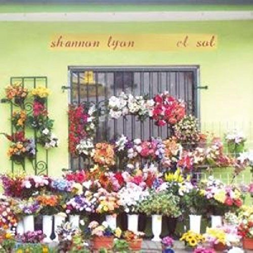 The image depicts a vibrant scene with a light green wall, adorned with numerous flowers in white pots, and a window featuring a black metal grate. The flowers are arranged on the windowsill and surrounding areas, creating a colorful display. Above the window, the text "shannon lyon el sol" is written in red cursive font on a yellow banner. Alt text: "Album cover with flowers in front of a green wall and window."