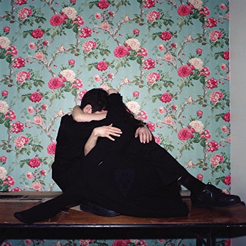 Two figures in black embrace against a floral wallpaper backdrop.