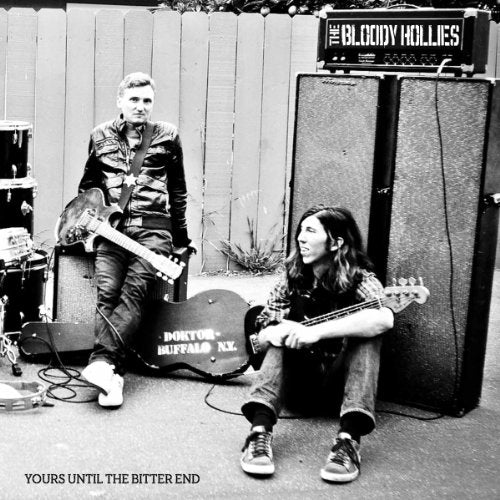 Black and white image of two musicians with guitars, seated near a large speaker.