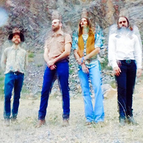 Four men with long hair and beards stand in front of a hillside.