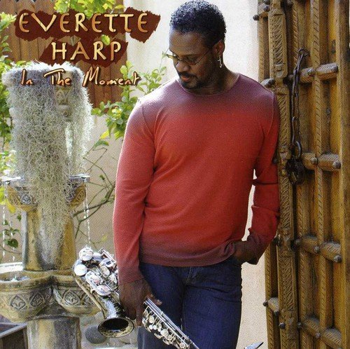 Man in red shirt holding saxophone stands beside ornate wooden door, outdoors.