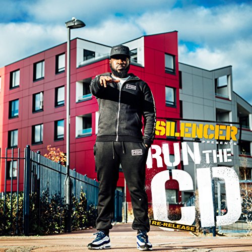 Man in black tracksuit stands before a red and gray building, pointing.