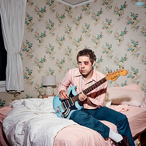 Man with a blue electric guitar sitting on a pink bed.