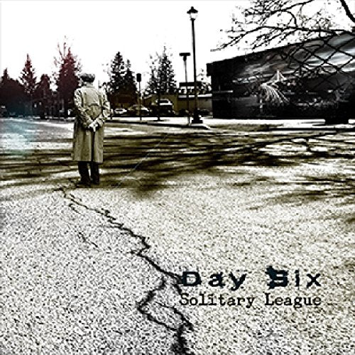 A man in a long coat stands alone on cracked asphalt, with a building and trees.
