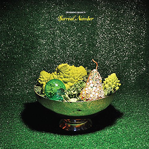 A surreal still life: a metal bowl holding pear, glass orb, and green plants.