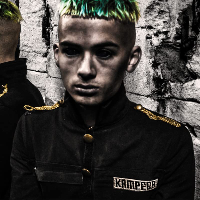 Young man with green hair in military-style jacket, looking intensely forward.