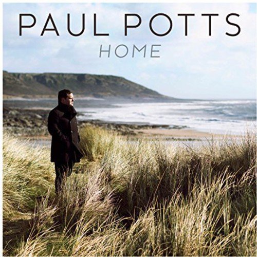 Man in dark coat stands in tall grass by a beach with a rocky shore.