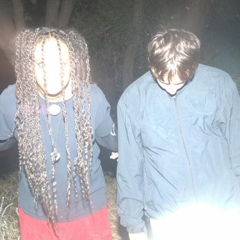Two people with long hair stand in a dimly lit, grassy area.