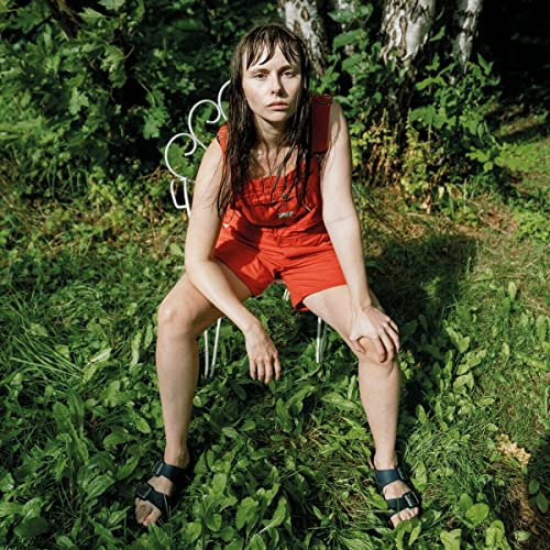 Woman in red overalls sits on a white chair in greenery.