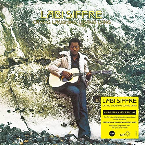 Labi Siffre sits with an acoustic guitar in front of a rocky waterfall.