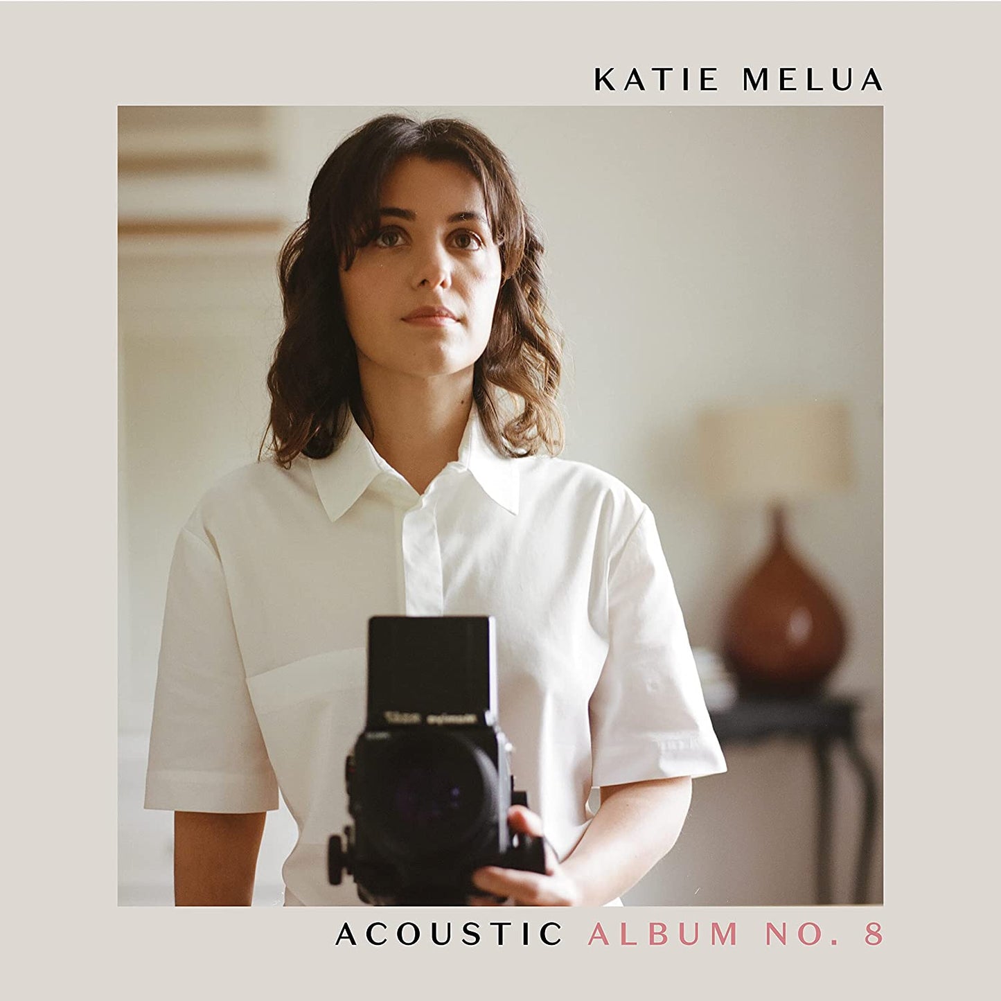 Katie Melua holding a camera, wearing a white shirt, on a beige album cover.