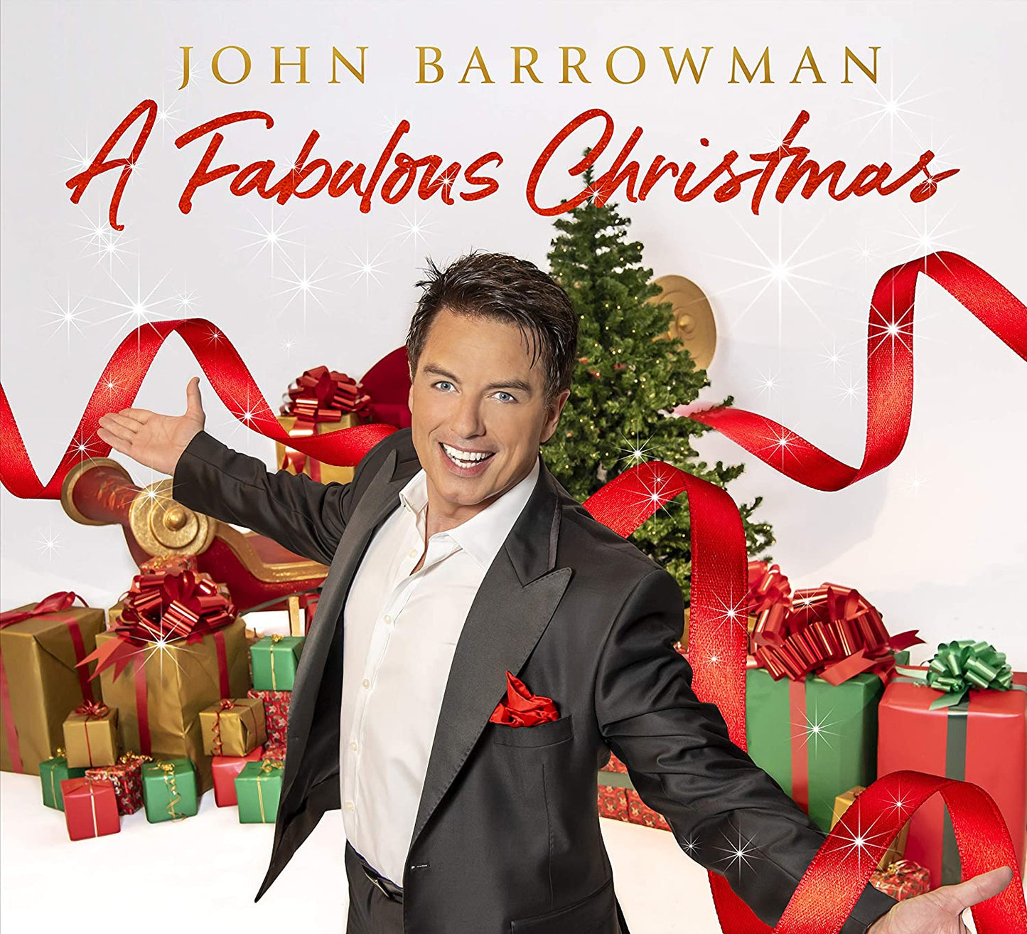 John Barrowman in a tuxedo, surrounded by Christmas gifts and a tree.